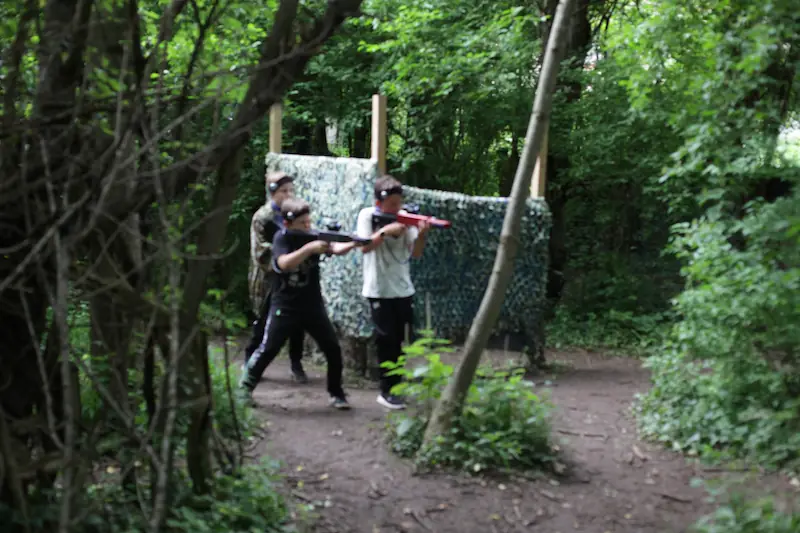 Lasertag Gruppen-Action und Taktik im Dschungel-Spielfeld Drei junge Spieler in taktischer Stellung hinter einer Tarnwand beim Outdoor Lasertag Wels. Ideal für Geburtstage, Gruppen und Schulklassen-Ausflüge.